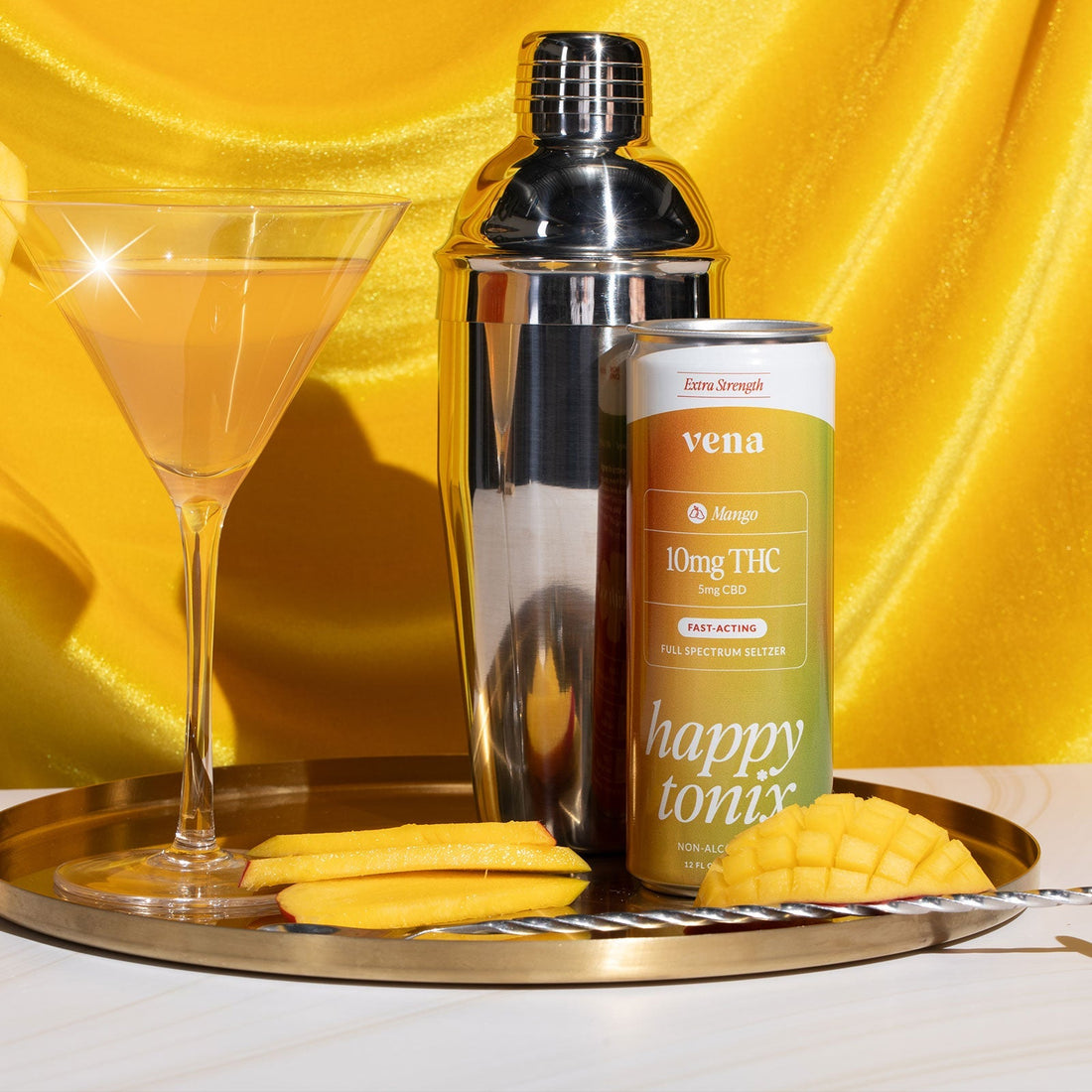 Close-up of Vena Mango 10mg THC seltzer can beside a martini glass, metal shaker, and mango slices on a yellow background