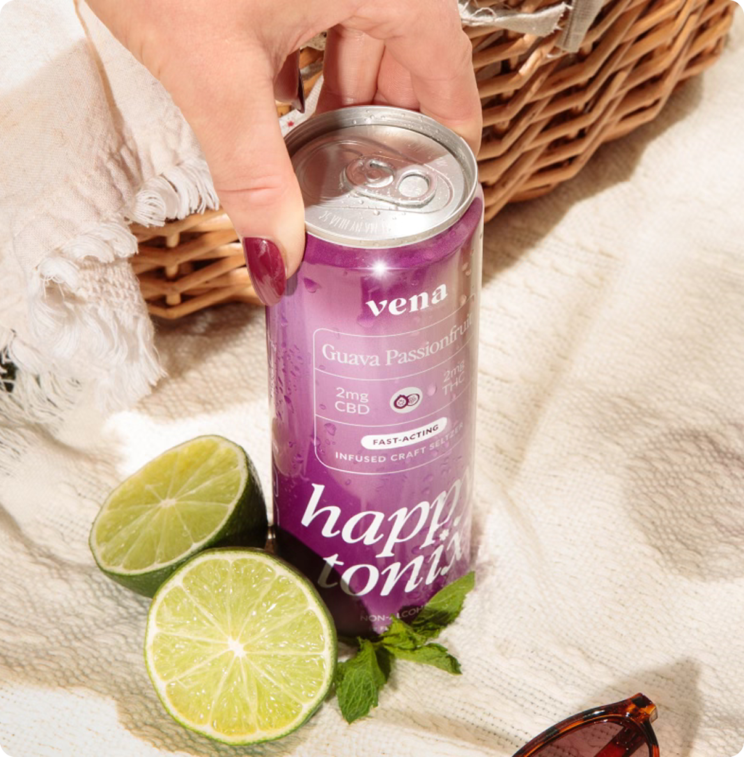 Top-down view of Vena Guava Passionfruit drink can showing 2mg CBD label, hand reaching for it on a picnic blanket with lime halves