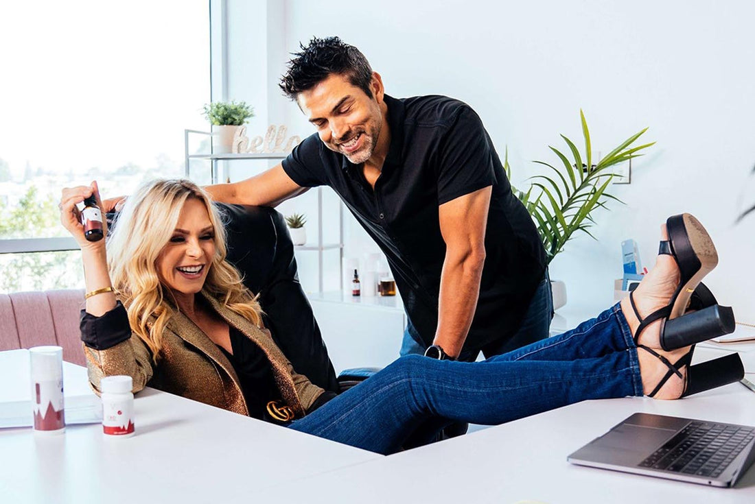 Smiling woman with feet on desk laughing while colleague leans in; office shelf shows hello sign and bottles.