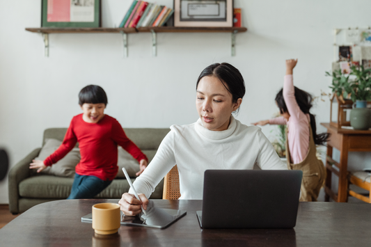Woman working on laptop and tablet at dining table while two children play in the background