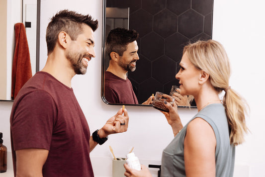 Smiling couple in bathroom: man holds a pill, woman holds a supplement bottle while they talk.