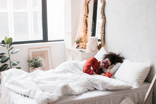 Woman in red pajamas sleeping with floral eye mask on a white bed, beside a mirror with string lights and a potted plant.