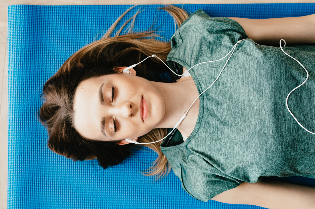 Young woman resting on a blue yoga mat with earbuds in, eyes closed, wearing a green shirt.