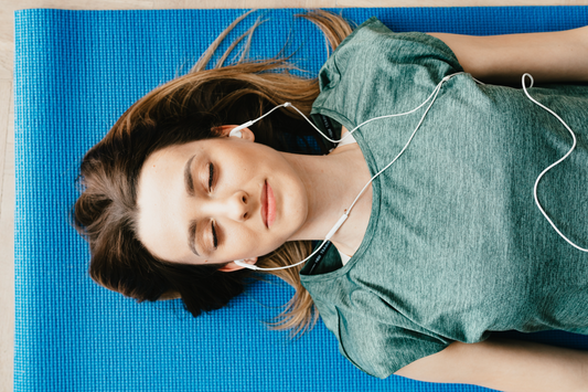 Young woman resting on a blue yoga mat with earbuds in, eyes closed, wearing a green shirt.