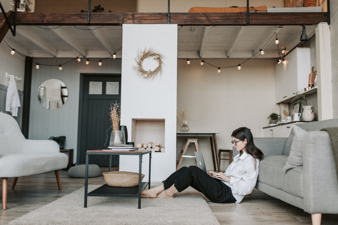 Woman sitting on living room floor using laptop beside sofa in cozy loft-style home office