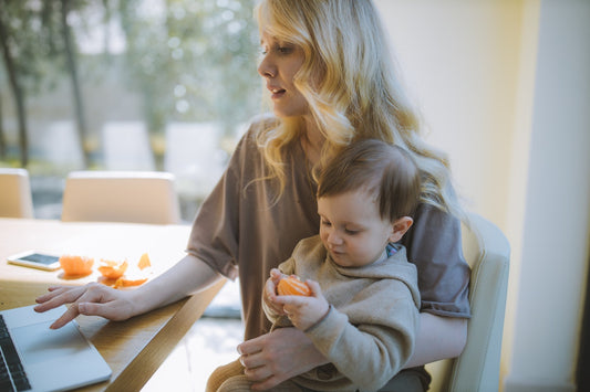 Mother holding a toddler on her lap while using a laptop at a dining table with peeled orange segments nearby.