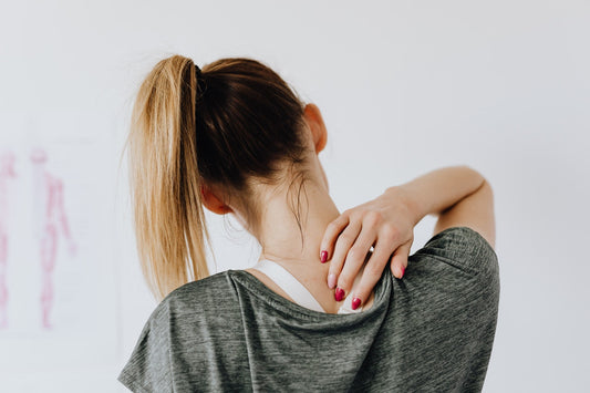 Woman seen from behind with ponytail holding her neck and shoulder, suggesting neck pain or muscle ache.
