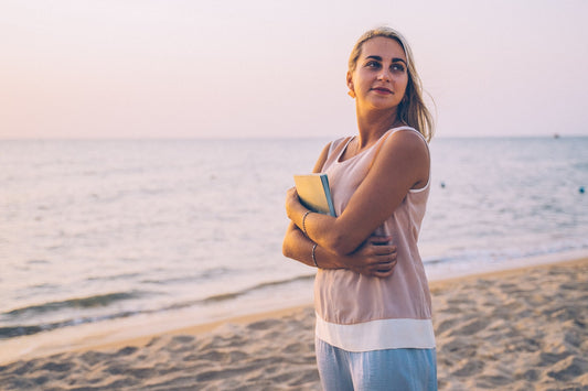 Woman standing on a beach at sunset holding a notebook, looking toward the horizon