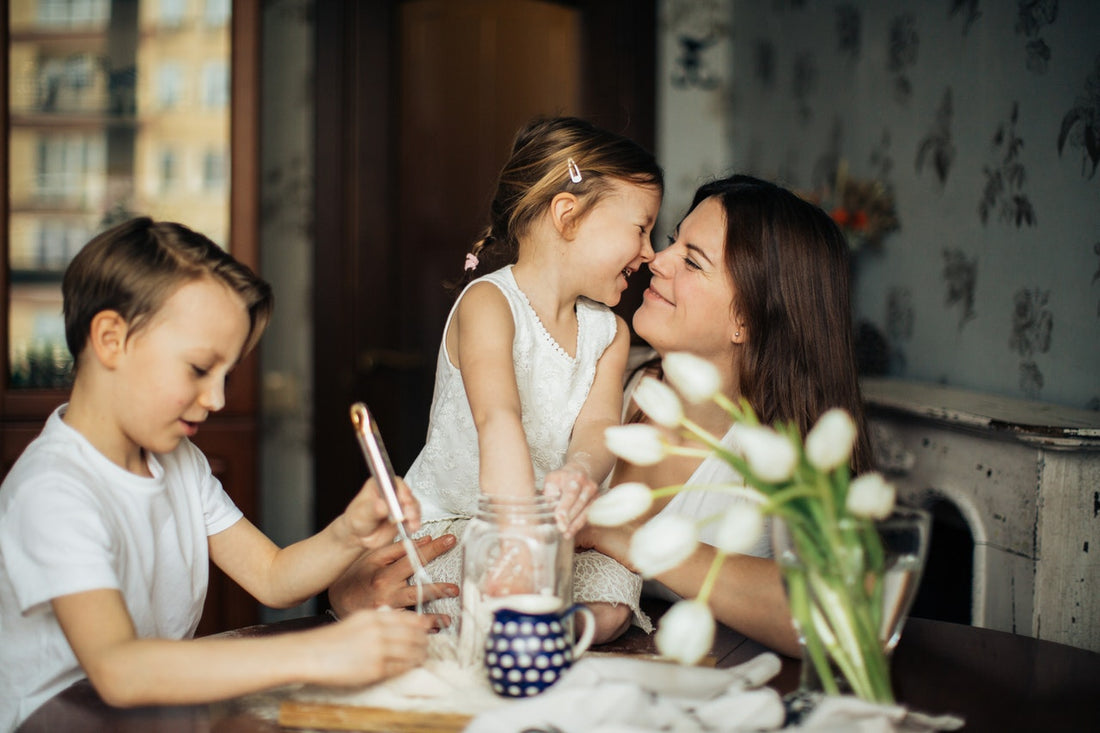 Mother smiling and holding daughter at a kitchen table while son stirs baking ingredients.