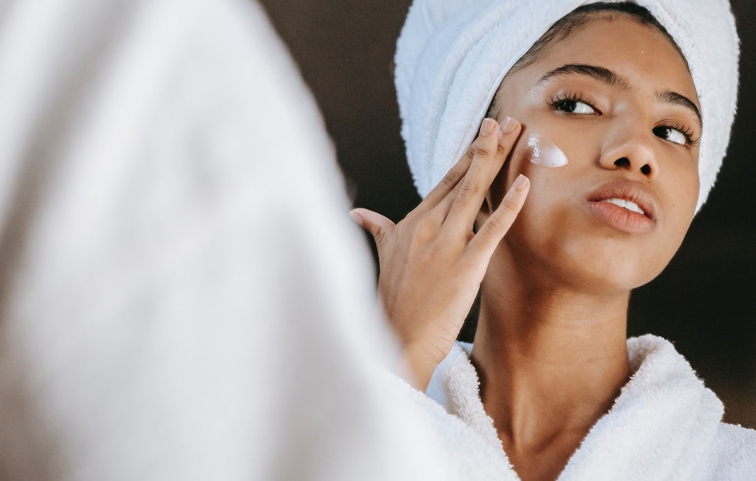 Young woman with towel on head applying cream to her cheek while looking in a mirror