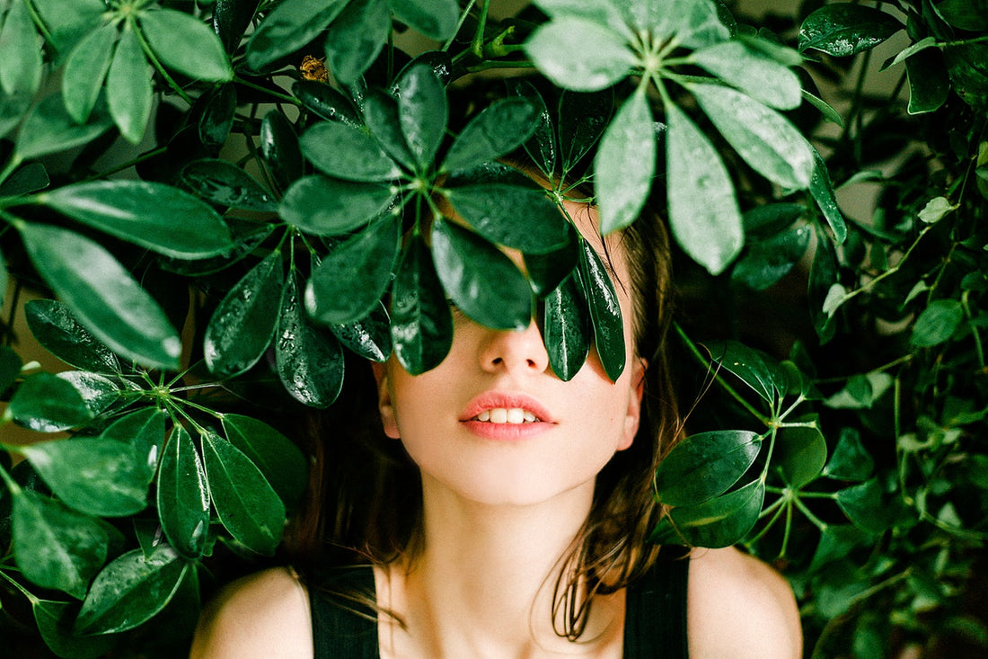 Close-up of a woman with green leaves covering her eyes, lips and chin visible among glossy tropical foliage.