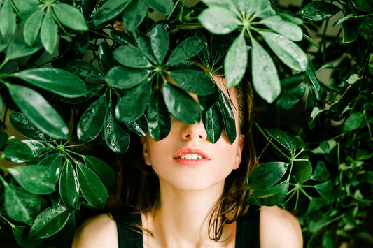 Close-up of a woman with green leaves covering her eyes, lips and chin visible among glossy tropical foliage.