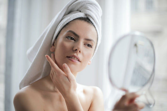Woman with towel on her head touching her cheek while looking into a handheld mirror
