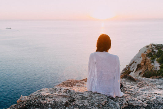 Person sitting on a rocky cliff facing the sea at sunset, back to camera