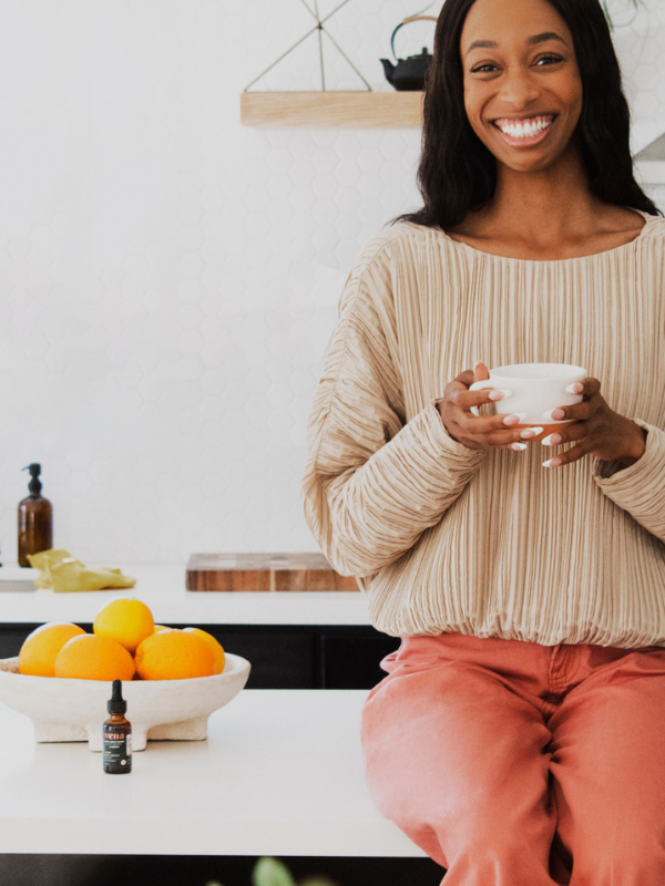 Smiling woman perched on a kitchen counter holding a cup, with a bowl of oranges and a small dropper bottle on the counter.