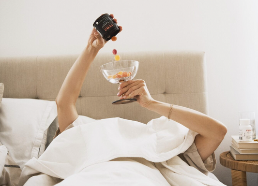 Person in bed pouring colorful gummy supplements from a jar labeled 'vena' into a glass dish