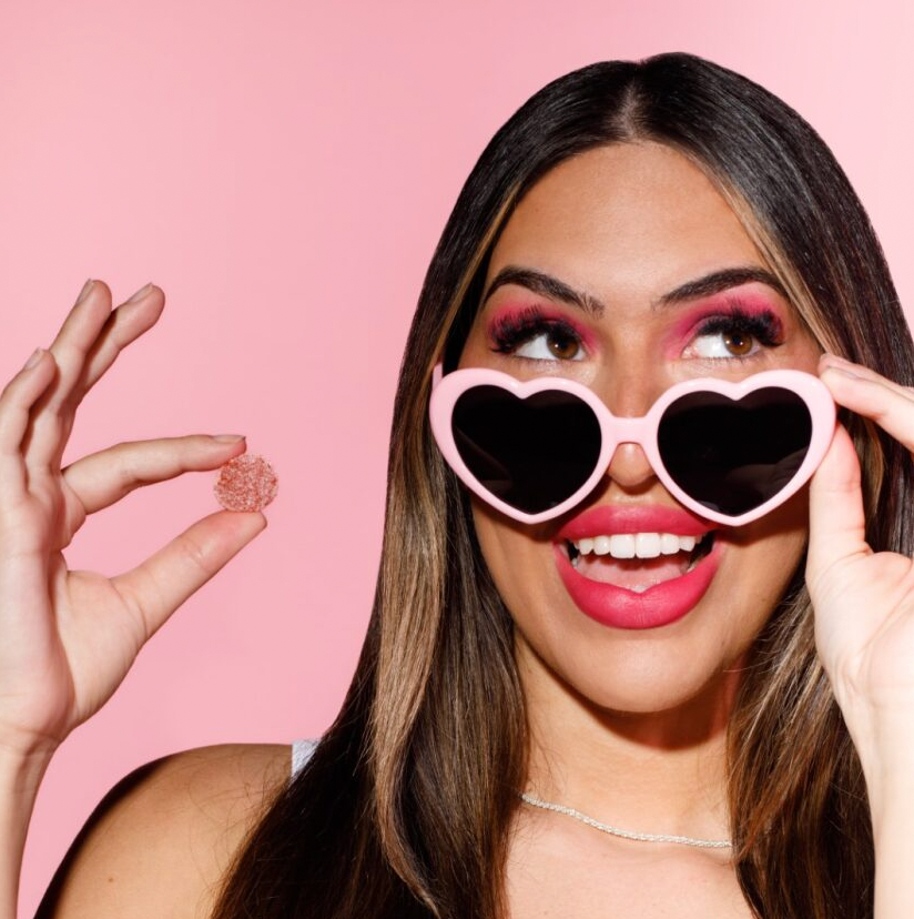 Smiling woman in heart-shaped pink sunglasses holding a pink gummy against a pink background