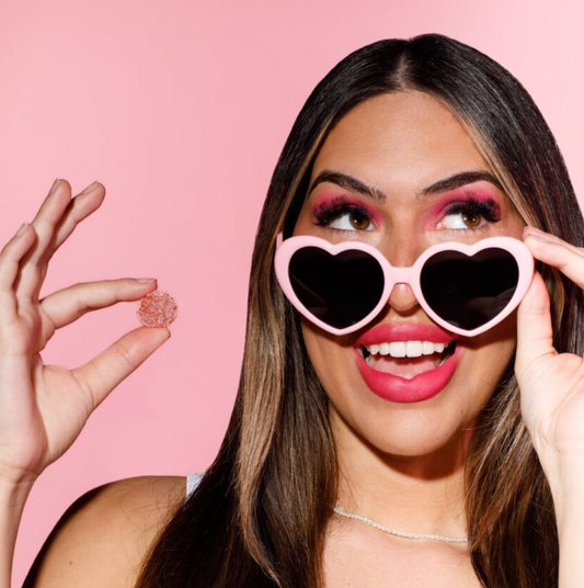 Smiling woman in heart-shaped pink sunglasses holding a pink gummy against a pink background
